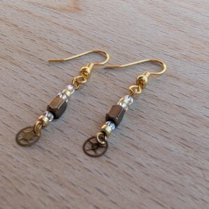May include: A pair of drop earrings featuring gold-tone ear wires, small clear beads, a central metallic bronze-colored cube bead, and a small circular gear-shaped charm at the bottom of each earring, creating a dangling design.