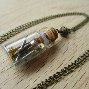 May include: A glass vial filled with small metal pieces hangs from a bronze chain necklace. The vial has a cork stopper and the chain has a decorative bronze charm.
