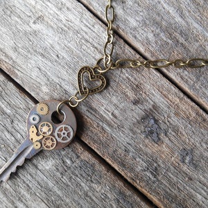 May include: A bronze key necklace with a heart charm and a key with gears and cogs attached. The necklace is on a wooden surface.