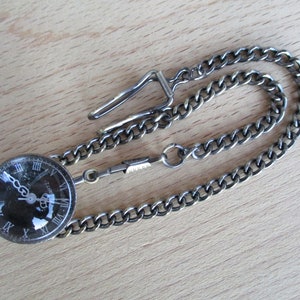 May include: A vintage pocket watch with a dark face and Roman numeral hour markers. The watch is attached to a silver-toned chain with a clasp. The watch and chain are displayed on a light wood surface.