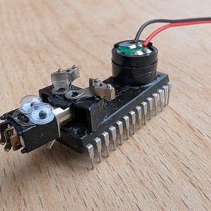 May include: A black electronic component featuring a row of silver metal pins along the side, a small circular black speaker with red and black wires attached, and two small white plastic eyes with black pupils mounted on the front section.