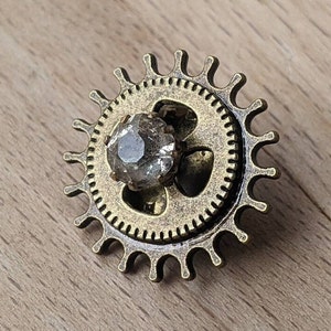 Pode incluir: Uma engrenagem de cor bronze com um cristal transparente no centro. A engrenagem tem um design decorativo e ornamentado com uma série de pontos ao redor da borda.