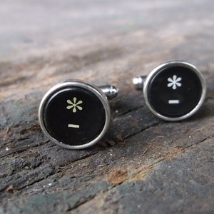 May include: A pair of silver cufflinks with black round buttons. Each button features a white asterisk and a horizontal line.