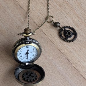 May include: A bronze-toned pocket watch pendant on a matching chain, featuring an open face with a white dial, black numerals, and quartz movement. The interior of the case displays a dark gear, and a separate gear charm hangs from the chain.