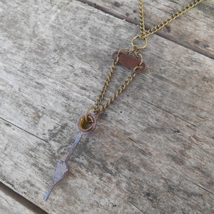 Peut inclure: Un collier en chaîne de laiton avec un accent en cuir marron et un long pendentif en métal bleu-gris fin.