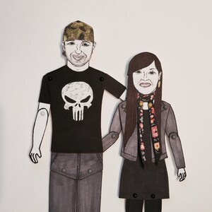 May include: A paper doll couple, the man wearing a black t-shirt with a white skull graphic and a camouflage hat, the woman wearing a gray jacket, a black dress, and a floral scarf.