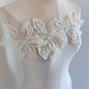 以下が含まれることがあります： 白いシアー生地に繊細な花のアップリケが施されています。アップリケは白いレースでできており、真珠のアクセントがついた大きな花が3つあります。