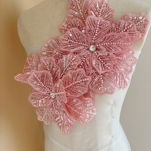 Può includere: Un'applicazione floreale rosa con paillettes e perline. L'applicazione è realizzata in tessuto delicato e presenta un design tridimensionale.