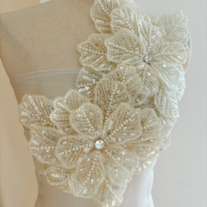 Puede incluir: Un par de grandes apliques de flores blancas con cuentas y centros de cristal. Los apliques están superpuestos y son perfectos para añadir un toque de elegancia a cualquier prenda.