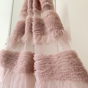 May include: A blush pink tulle curtain with multiple layers of ruffles. The curtain is sheer and delicate, with a soft, romantic look.