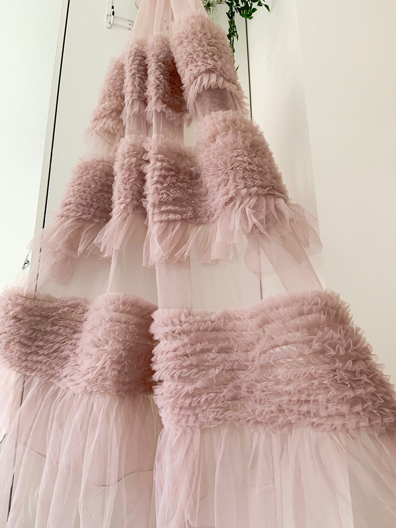 May include: A blush pink tulle curtain with multiple layers of ruffles. The curtain is sheer and delicate, with a soft, romantic look.