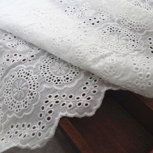 May include: White cotton eyelet lace trim with a scalloped edge and a delicate floral pattern. The lace is folded over itself, showing the intricate details of the design.
