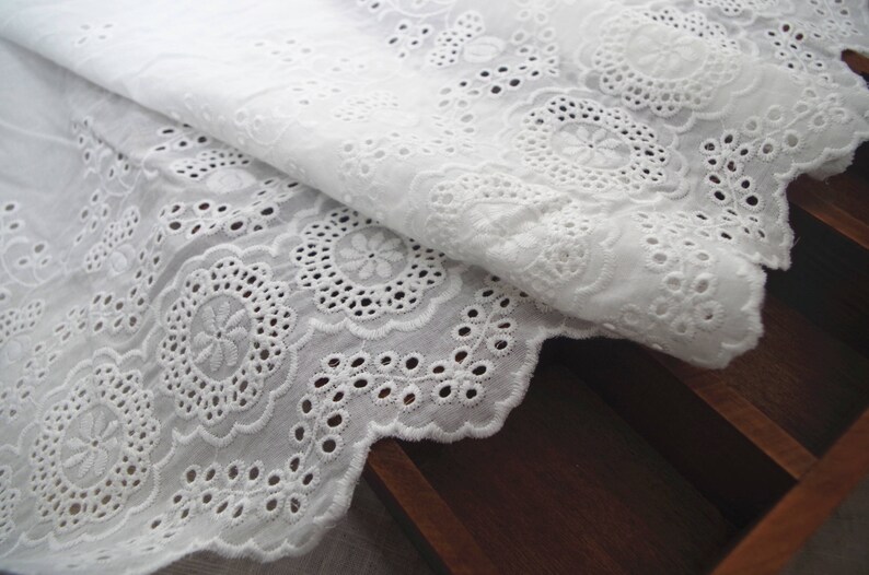 May include: White cotton eyelet lace trim with a scalloped edge and a delicate floral pattern. The lace is folded over itself, showing the intricate details of the design.
