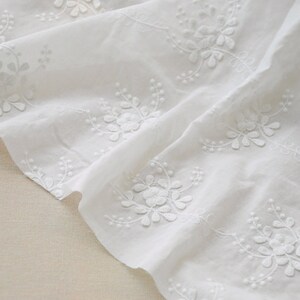May include: White cotton fabric featuring delicate embroidered floral designs. The fabric is folded and draped, highlighting the intricate details of the embroidery.