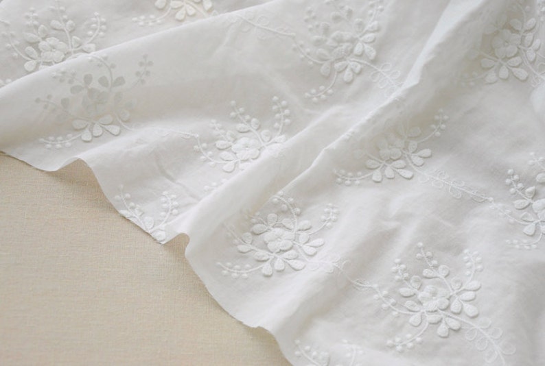 May include: White cotton fabric featuring delicate embroidered floral designs. The fabric is folded and draped, highlighting the intricate details of the embroidery.