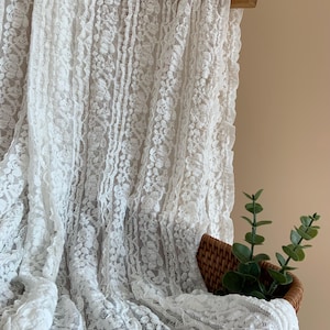 May include: White lace fabric with a floral design, hanging from a wooden rod. The delicate material is draped over a small woven basket containing green plant stems.