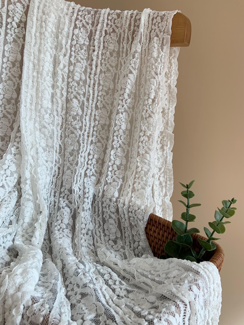 May include: White lace fabric with a floral design, hanging from a wooden rod. The delicate material is draped over a small woven basket containing green plant stems.