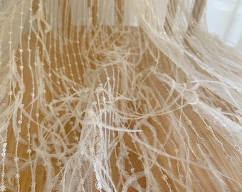Puede incluir: Tela de tul blanco con delicados detalles de plumas y pequeñas lentejuelas. La tela tiene una textura suave y fluida, ideal para crear prendas o accesorios elegantes.