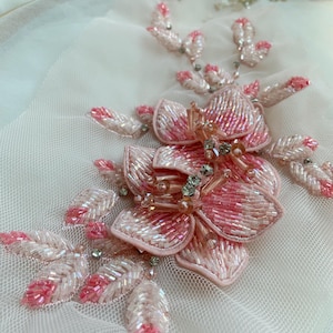 Puede incluir: Una aplicación floral sobre tela de malla blanca transparente, con pétalos superpuestos bordados con cuentas de vidrio rosas y blancas. El diseño incluye delicadas hojas con cuentas que se extienden desde la flor central con cristales.