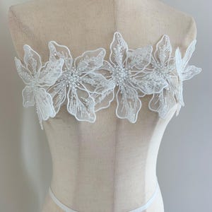 May include: White floral lace applique with beaded centres. The applique features five large, detailed flowers with delicate, openwork petals. The lace is attached to a mannequin, showcasing its potential use in garment design or embellishment.
