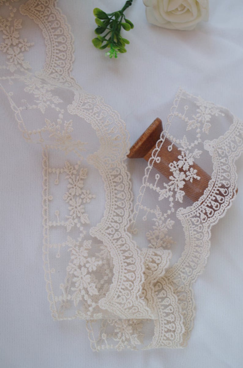 May include: A spool of ivory-coloured lace trim with a delicate floral pattern. The lace is soft and sheer, with a scalloped edge.