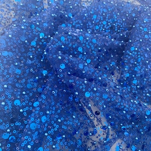 Puede incluir: Tela azul con lentejuelas pequeñas y grandes. Las lentejuelas están dispersas aleatoriamente por la tela.