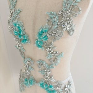 May include: A delicate, silver-toned lace applique with turquoise embellishments. The applique features a floral design with leaves and flowers, and is adorned with sparkling rhinestones.