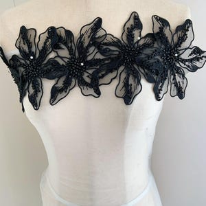 Op de afbeelding: Zwarte bloemen kanten applicatie met pailletten en kraal versieringen. De applicatie heeft drie grote bloemmotieven met delicate, doorschijnende bloemblaadjes. Het midden van elke bloem is dicht versierd met pailletten en kralen.