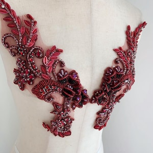 May include: Two ornate, red beaded appliques with floral and leaf designs. The appliques are embellished with sequins and beads, creating a textured and dimensional effect. The design is symmetrical, with each piece mirroring the other.