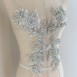 May include: A silver beaded and lace applique with floral designs. The applique is ideal for adding a touch of sparkle and elegance to any garment.