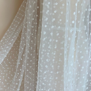 May include: White floral embroidered tulle fabric with a delicate pattern of small white flowers. The fabric is sheer and lightweight, making it ideal for crafting and sewing projects.