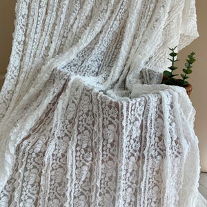 May include: White lace fabric with a floral design. The fabric is draped over a chair, creating soft folds and shadows. The lace has a delicate, textured appearance, and the overall effect is elegant and romantic.