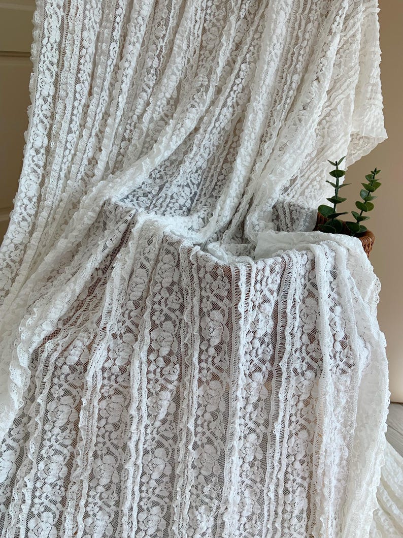 May include: White lace fabric with a floral design. The fabric is draped over a chair, creating soft folds and shadows. The lace has a delicate, textured appearance, and the overall effect is elegant and romantic.