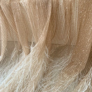 K&ouml;nnte beinhalten: Beigefarbener Stoff mit schimmernder, paillettenbesetzter Textur und einem wei&szlig;en Federbesatz. Der Stoff ist drapiert und gefaltet, wodurch die Textur und der Federbesatz sichtbar werden.