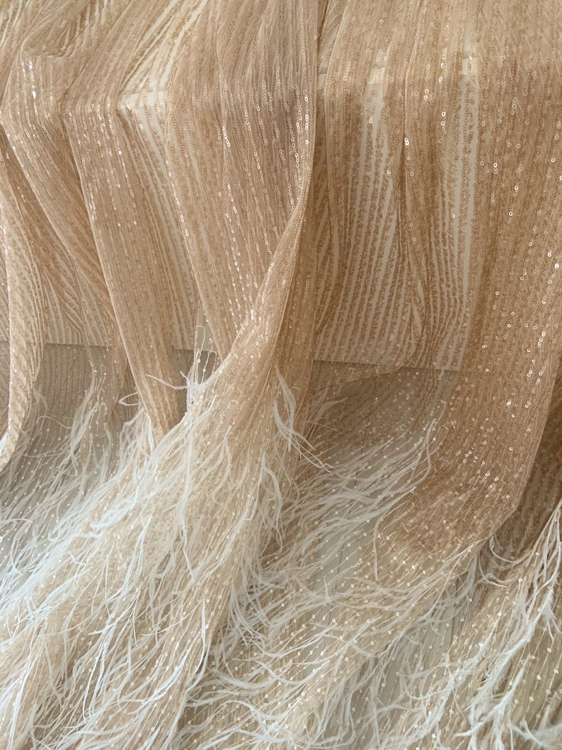 K&ouml;nnte beinhalten: Beigefarbener Stoff mit schimmernder, paillettenbesetzter Textur und einem wei&szlig;en Federbesatz. Der Stoff ist drapiert und gefaltet, wodurch die Textur und der Federbesatz sichtbar werden.