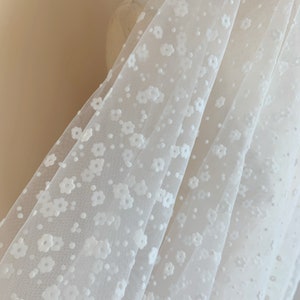 May include: White tulle fabric with a delicate floral pattern. The fabric is sheer and has a soft, romantic feel.