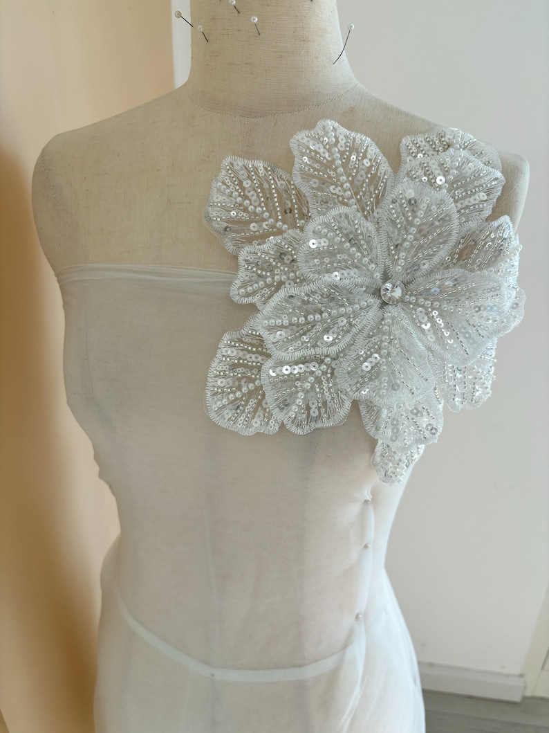 Pu&ograve; includere: Un tessuto bianco trasparente con una grande applicazione floreale bianca perlata. Il fiore ha un strass centrale ed &egrave; fissato al tessuto con spilli.