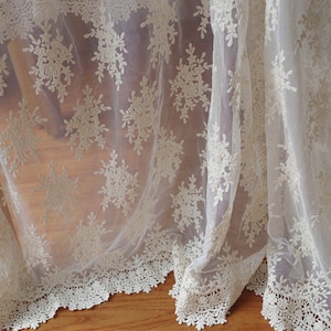 May include: Ivory-colored lace fabric with a delicate floral pattern. The sheer material drapes gracefully, showcasing intricate embroidered details. The bottom edge features a scalloped lace trim, adding a touch of elegance. Ideal for curtains or decorative accents.
