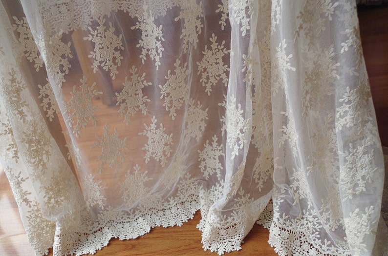 May include: Ivory-colored lace fabric with a delicate floral pattern. The sheer material drapes gracefully, showcasing intricate embroidered details. The bottom edge features a scalloped lace trim, adding a touch of elegance. Ideal for curtains or decorative accents.