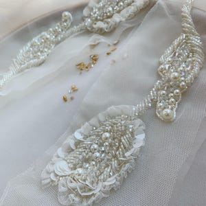Puede incluir: Dos aplicaciones de tela de tul blanco con bordados intrincados de cuentas de vidrio transparente, perlas blancas y lentejuelas iridiscentes. El diseño incluye formas alargadas de hojas y motivos florales, con cuentas sueltas sobre la tela.