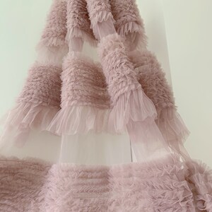 May include: A pale pink tulle fabric with multiple layers of ruffles. The fabric is soft and delicate, with a flowing, ethereal look.