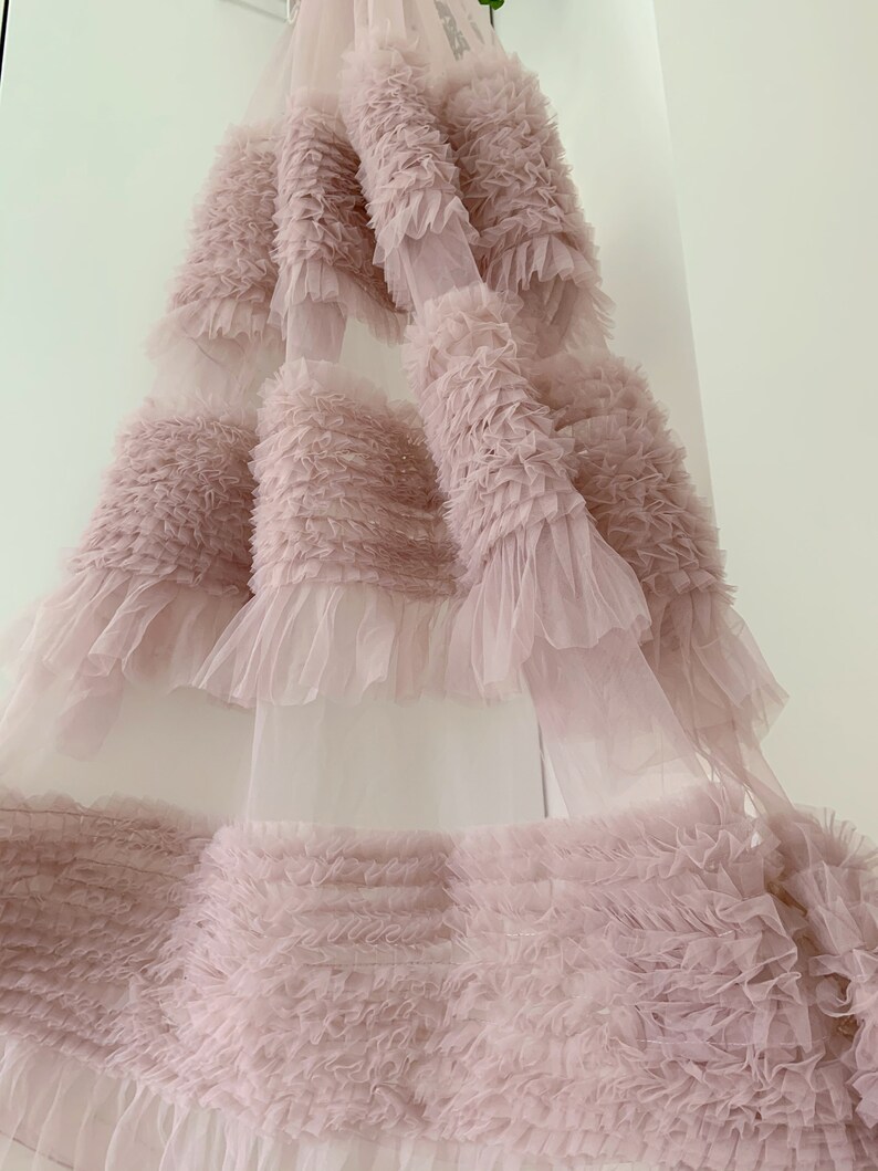 May include: A pale pink tulle fabric with multiple layers of ruffles. The fabric is soft and delicate, with a flowing, ethereal look.