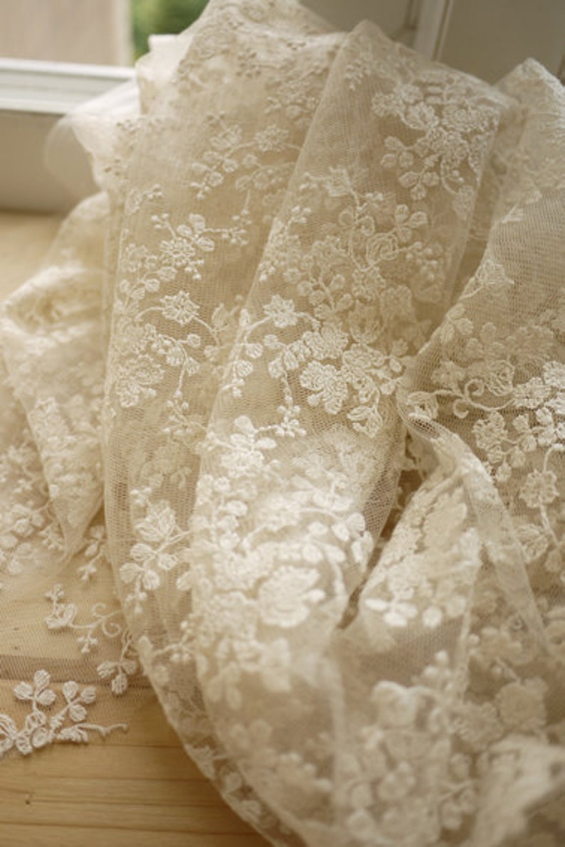 Puede incluir: Tela de encaje blanco marfil con un delicado patr&oacute;n floral. El encaje es transparente y tiene una textura suave y fluida.