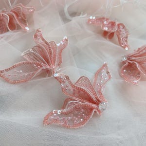 May include: Several three-dimensional floral appliques in a soft blush pink hue, crafted from sheer mesh fabric and embellished with matching sequins and beads. Each flower features layered, ruffled petals with a delicate, translucent appearance.
