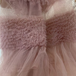 May include: A soft, pink tulle fabric with a ruffled texture. The fabric is layered and has a delicate, airy feel.