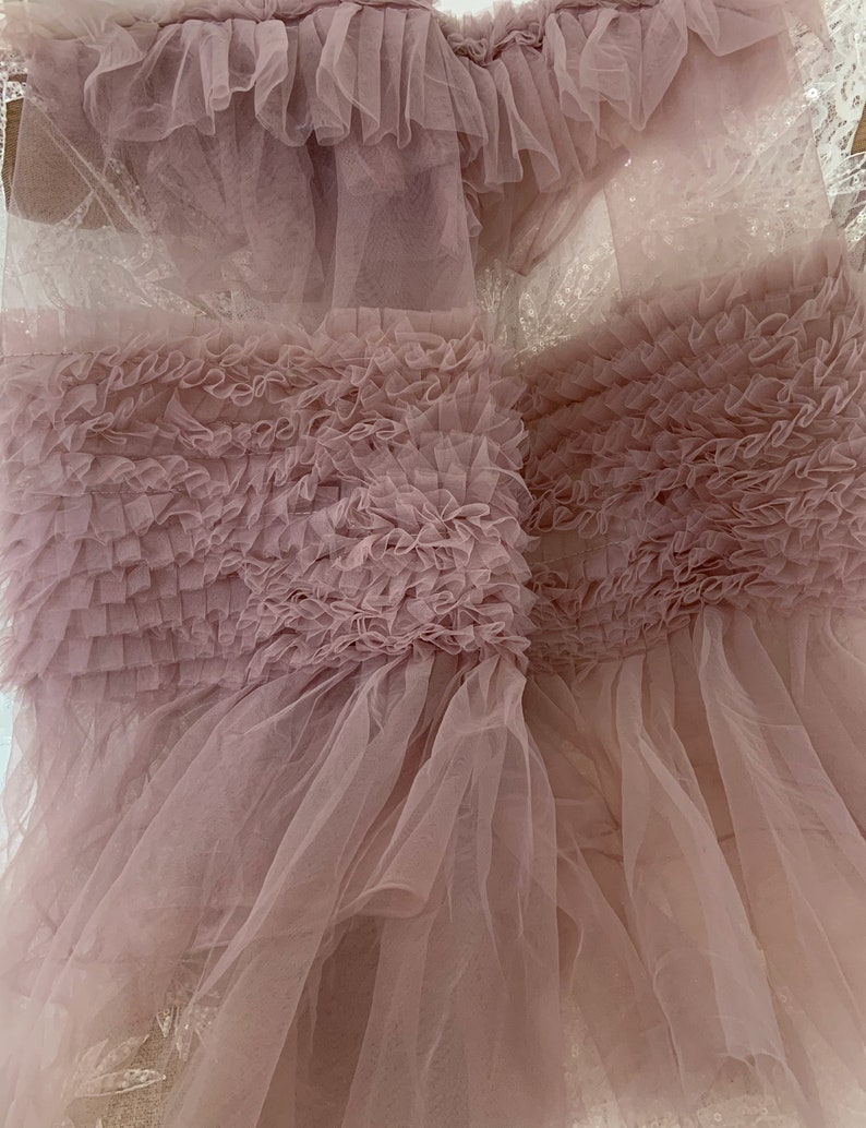 May include: A soft, pink tulle fabric with a ruffled texture. The fabric is layered and has a delicate, airy feel.