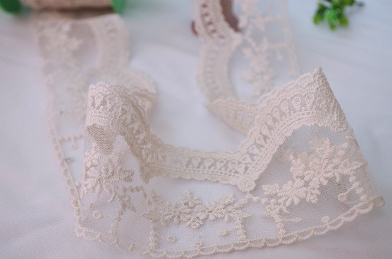 May include: A close-up of a delicate, off-white lace trim with a floral pattern. The lace is soft and sheer, with a delicate texture. It is perfect for adding a touch of elegance to any project.