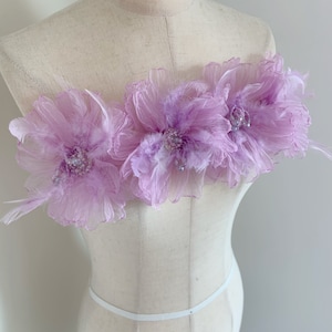 May include: Three large, light purple fabric flowers with beaded centres, arranged horizontally on a white dress form.
