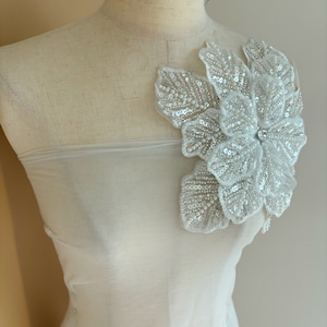 Pu&ograve; includere: Un tessuto bianco trasparente con una grande applicazione di fiore bianco perlato. Il fiore ha un centro di perla ed &egrave; fatto di paillettes e perline.
