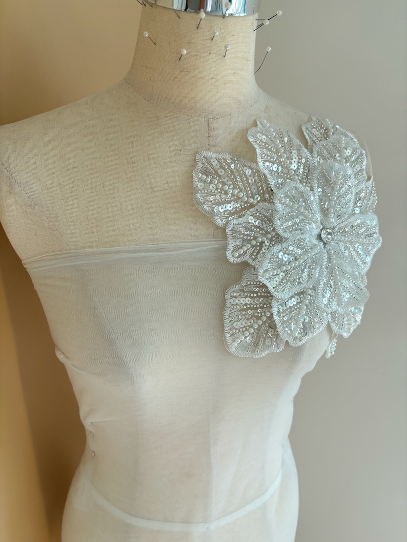 Pu&ograve; includere: Un tessuto bianco trasparente con una grande applicazione di fiore bianco perlato. Il fiore ha un centro di perla ed &egrave; fatto di paillettes e perline.